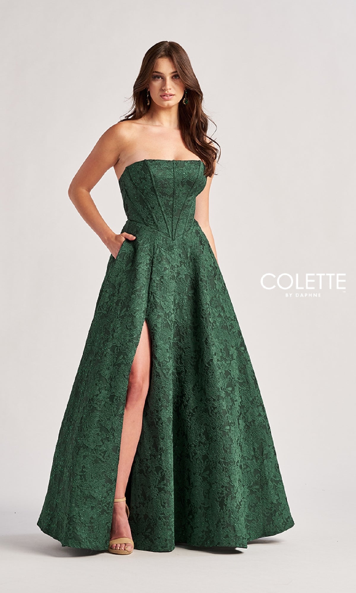 A woman models the Colette CL8675 long prom dress, featuring a strapless neckline, green floral a-line skirt, floor-length hem, front slit, and pockets as she poses against a plain background.